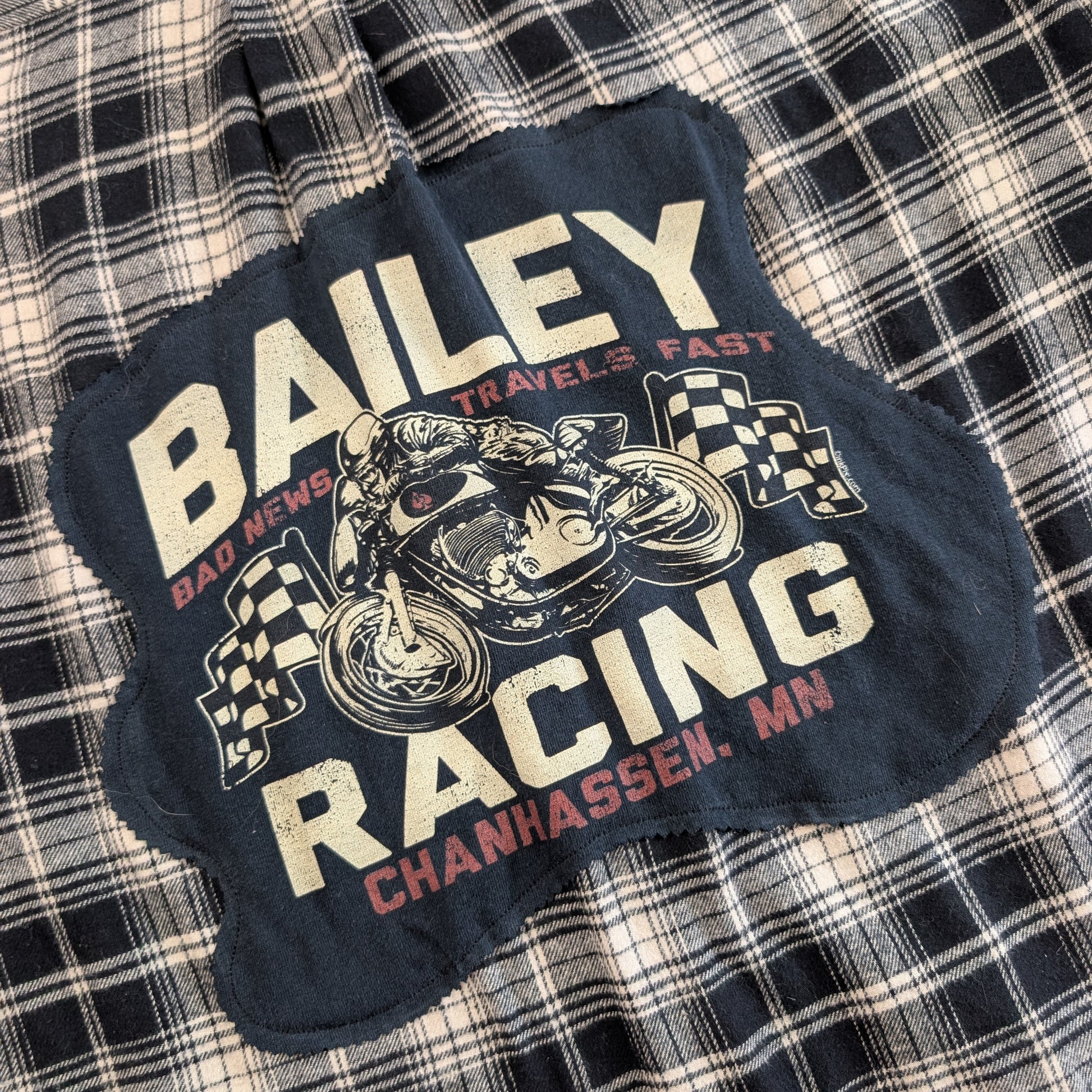 A dark navy blue graphic patch featuring a vintage motorcycle racer and the text 'BAILEY RACING CHANHASSEN, MN' in cream and red, laid flat on a black and white plaid fabric background.
