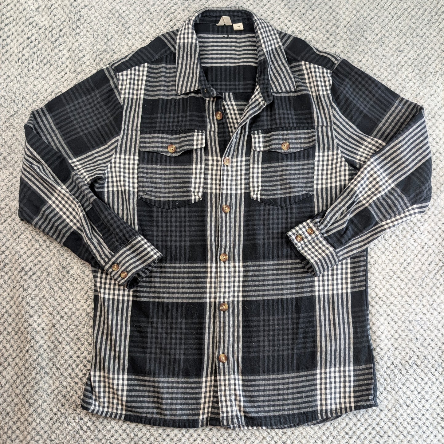 A long-sleeve plaid flannel shirt laid flat on a textured gray carpet. The shirt features a black, gray, and white plaid pattern with brown buttons.