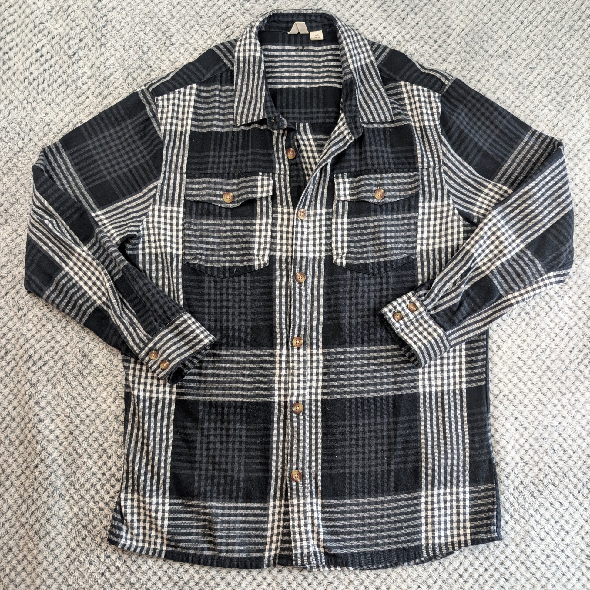 A long-sleeve plaid flannel shirt laid flat on a textured gray carpet. The shirt features a black, gray, and white plaid pattern with brown buttons.