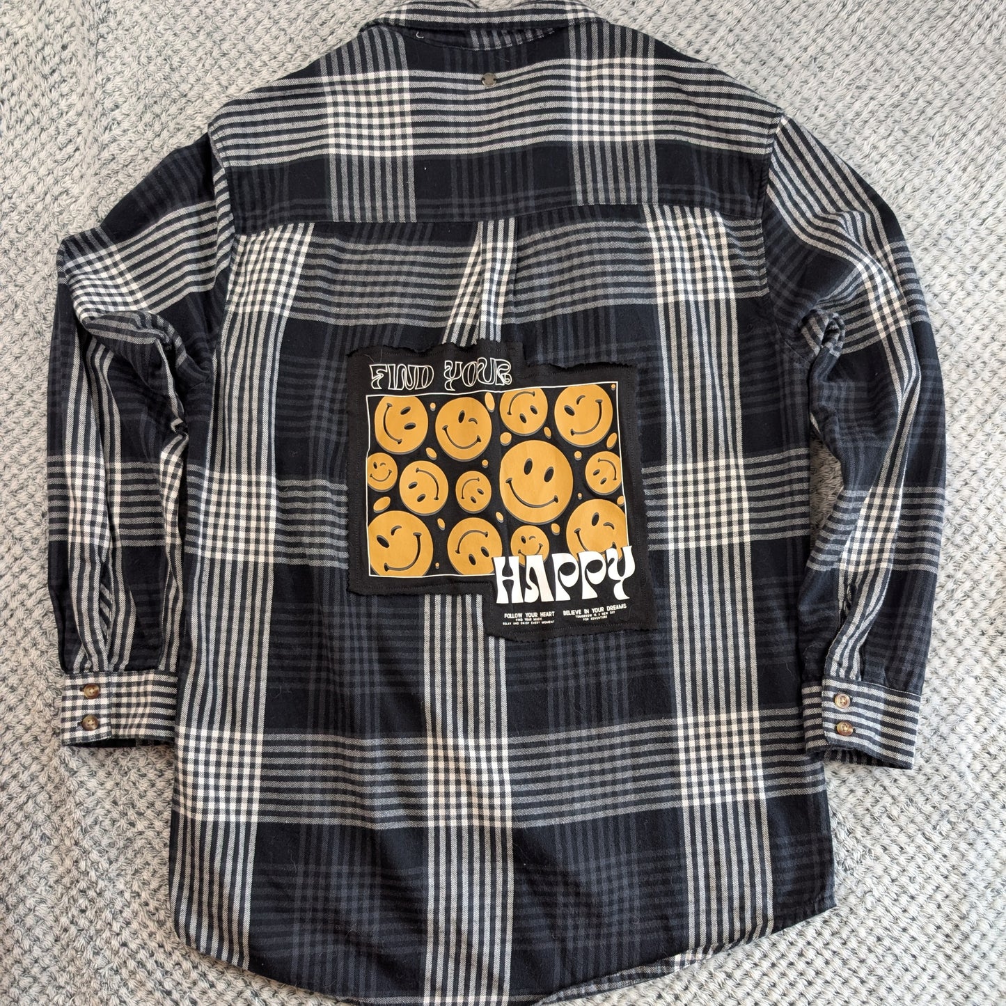 A black and white plaid flannel shirt laid flat on a textured gray carpet, featuring a large graphic patch on the back with yellow smiley faces and the word 'HAPPY'.