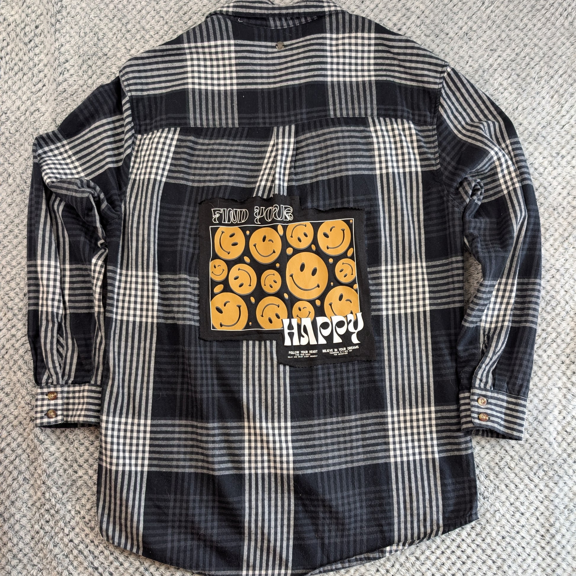 A black and white plaid flannel shirt laid flat on a textured gray carpet, featuring a large graphic patch on the back with yellow smiley faces and the word 'HAPPY'.