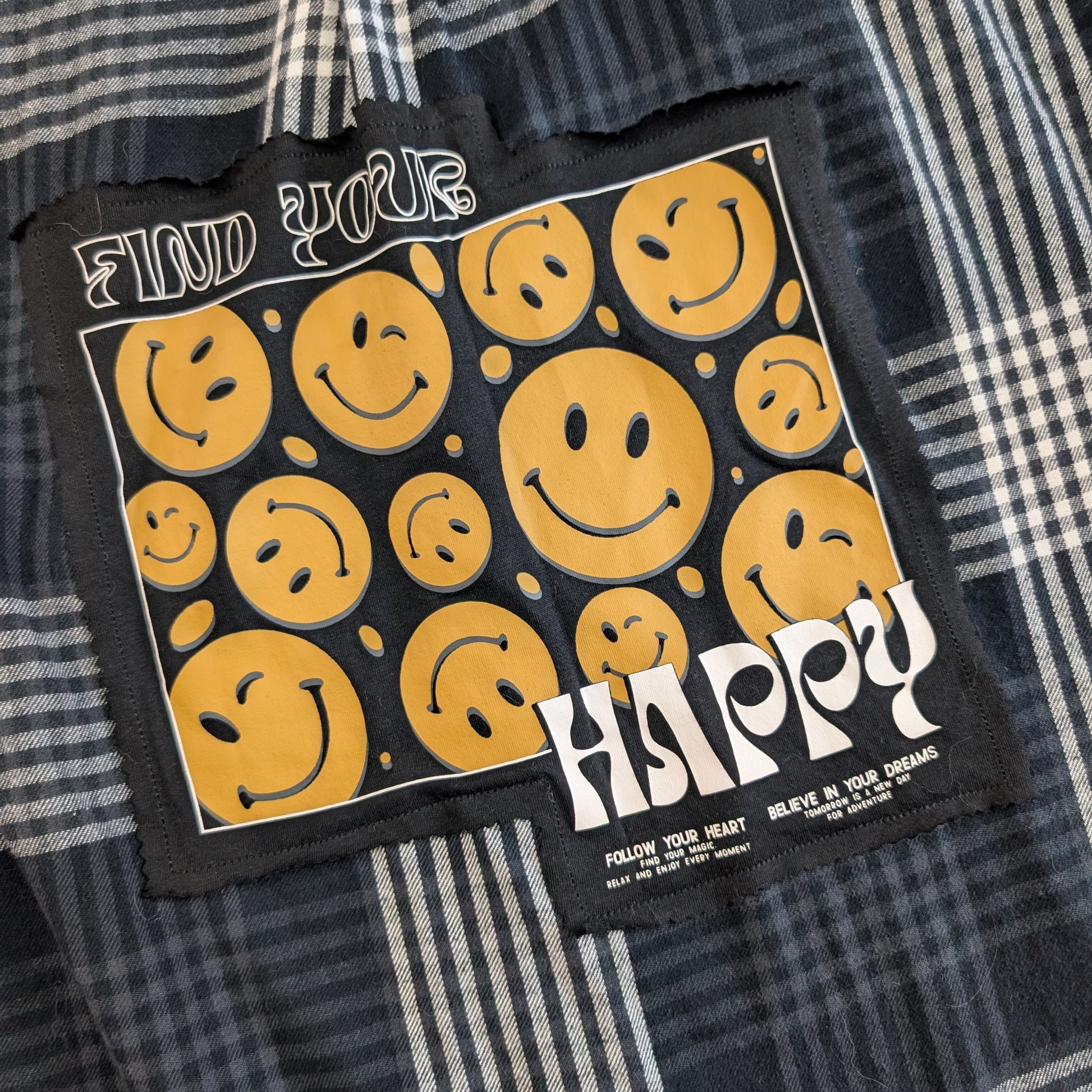 A square black embroidered patch featuring yellow smiley faces and the words 'FIND YOUR HAPPY' in white, laid flat on a plaid shirt.