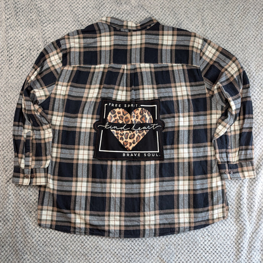 A long-sleeve plaid flannel shirt laid flat on a textured gray blanket. The shirt features a navy, beige, and gray plaid pattern with a central graphic patch that reads 'Free Spirit.