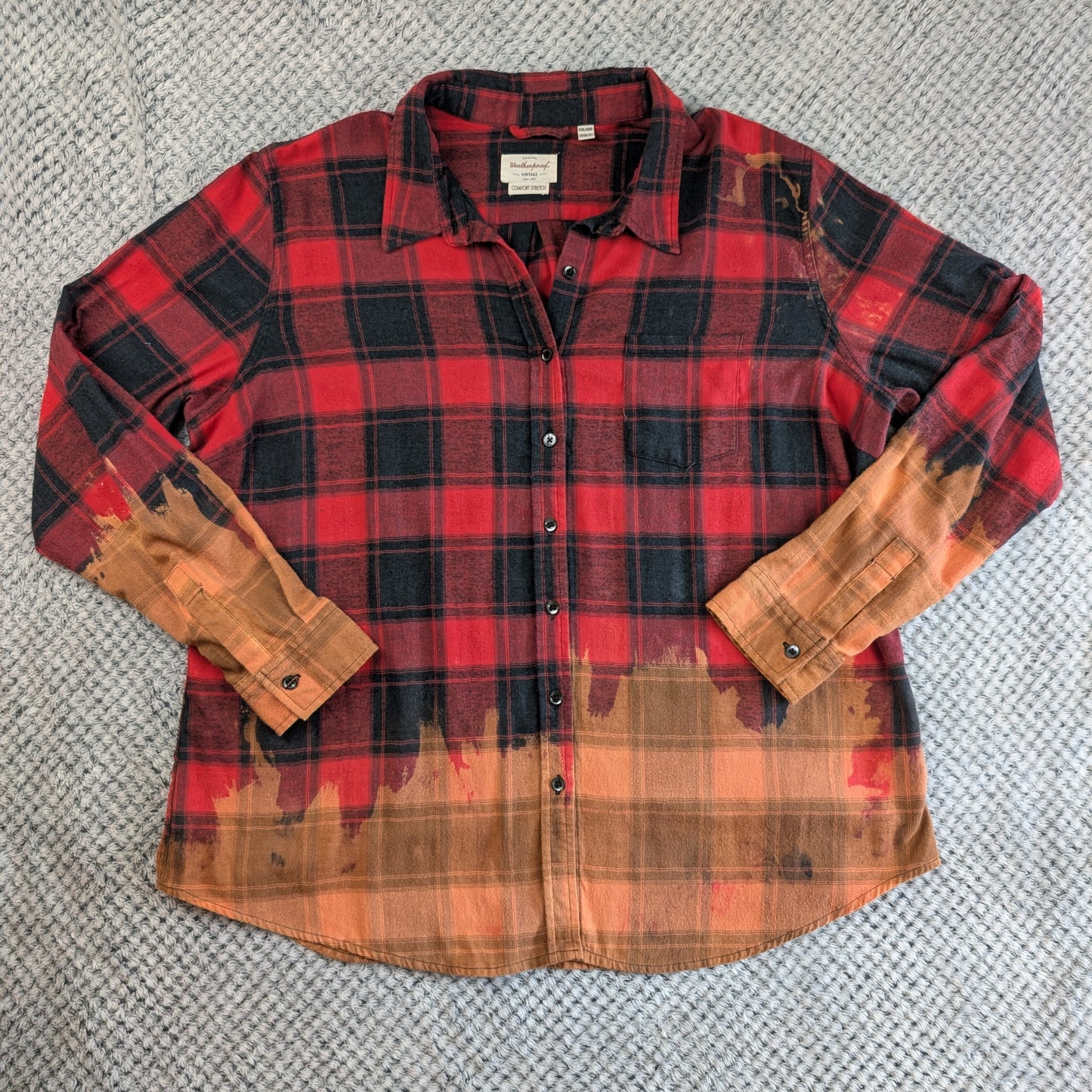 A long-sleeve button-up flannel shirt laid flat on a gray textured carpet. The shirt features a red and black plaid pattern on the upper body and sleeves, fading into a distressed orange and brown pattern at the hem and cuffs.