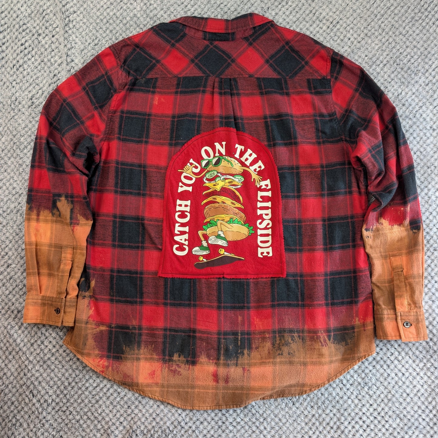 A red and black plaid long-sleeve shirt laid flat on a gray textured carpet, featuring a large red back patch with a taco illustration and the words 'CATCH YOU ON THE LIPSIDE'.