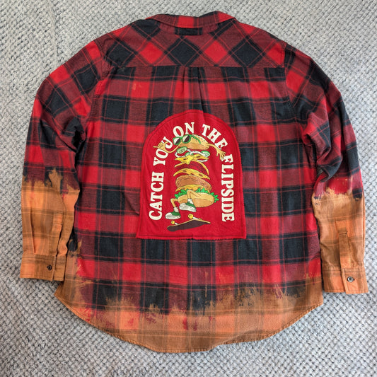 A red and black plaid long-sleeve shirt laid flat on a gray textured carpet, featuring a large red back patch with a taco illustration and the words 'CATCH YOU ON THE LIPSIDE'.
