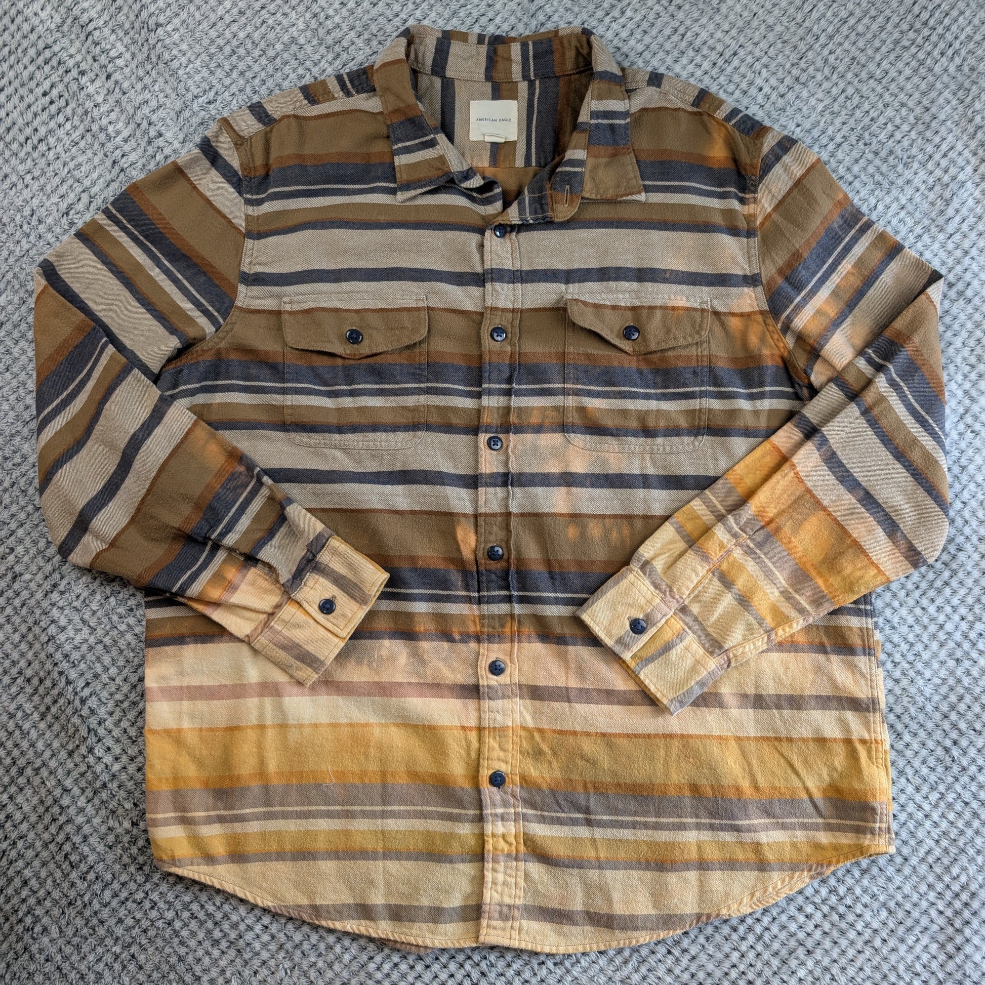 A long-sleeve button-up shirt with horizontal stripes in brown, beige, and muted blue-gray, laid flat on a gray textured fabric background.