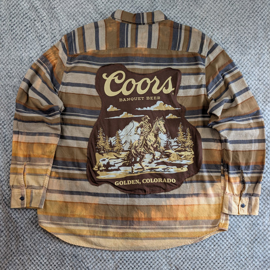 A long-sleeve striped flannel shirt laid flat on a gray textured surface, featuring a large Coors Banquet Beer Golden, Colorado graphic patch on the back.
