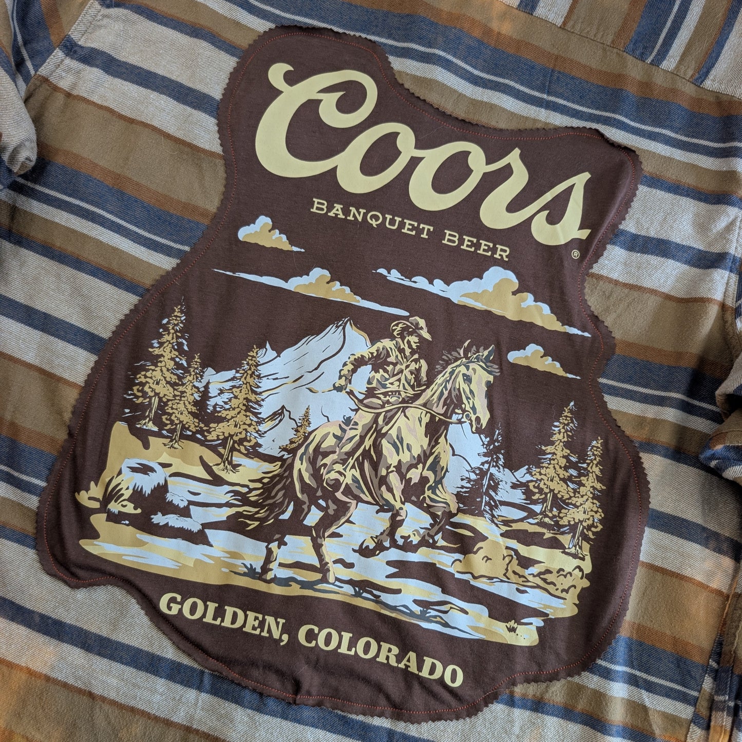 A Coors Banquet Beer graphic patch featuring a cowboy on a horse in a mountain landscape, laid flat on a blue and tan striped shirt.