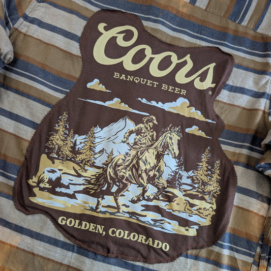 A Coors Banquet Beer graphic patch featuring a cowboy on a horse in a mountain landscape, laid flat on a blue and tan striped shirt.
