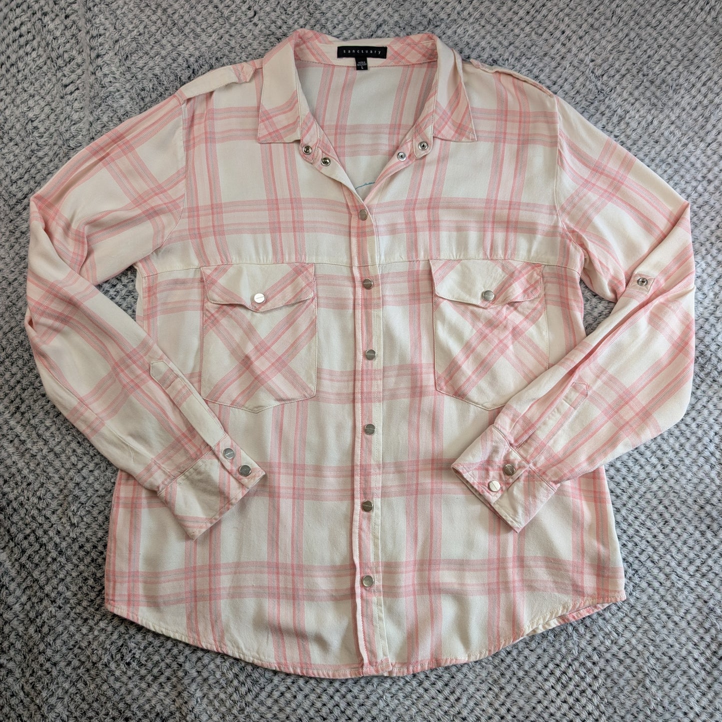 A long-sleeve plaid button-up shirt laid flat on a gray textured carpet. The shirt features a cream base with a pink plaid pattern, two chest pockets, and silver-toned snap buttons.