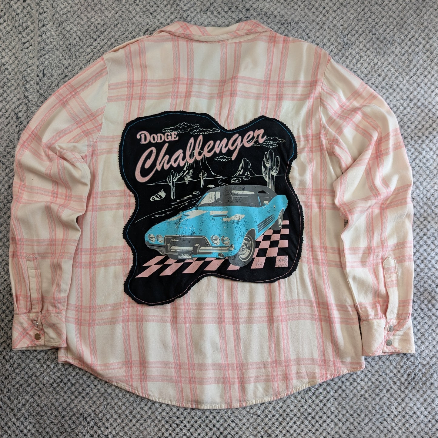 A pink and cream plaid flannel shirt laid flat on a gray textured carpet, showing a large back patch with a vintage blue Dodge Challenger car and the words 'Dodge Challenger'.