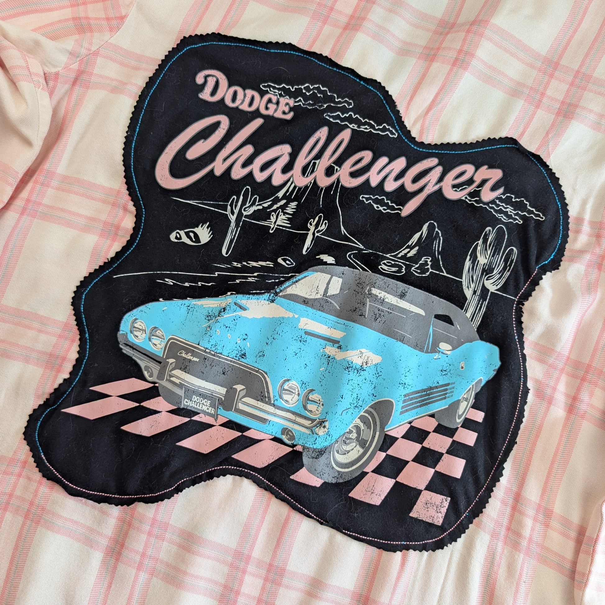 A close-up of a black fabric patch featuring a distressed light blue Dodge Challenger car, cacti, mountains, and the words 'Dodge Challenger' in pink and white, laid flat on a pink and white plaid fabric background.