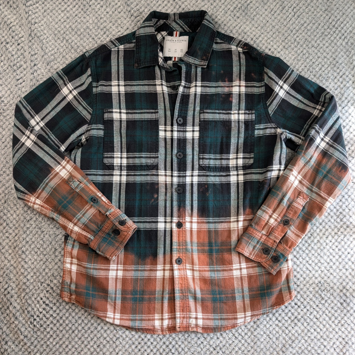 A long-sleeve plaid button-up shirt laid flat on a textured gray woven surface. The shirt features a dark green and black plaid upper body with white stripes, transitioning into a rust-orange and brown plaid pattern on the lower body and sleeves.