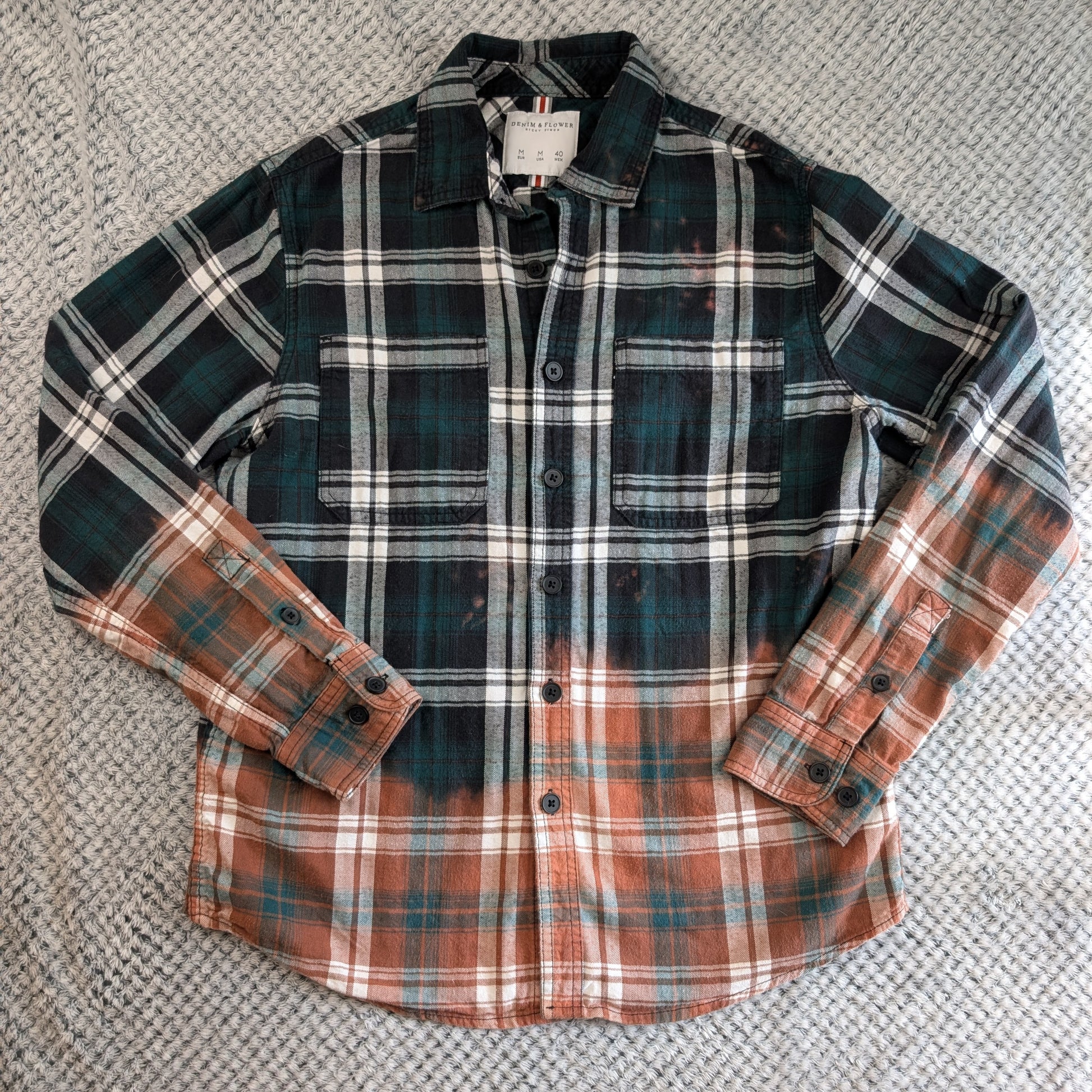 A long-sleeve plaid button-up shirt laid flat on a textured gray woven surface. The shirt features a dark green and black plaid upper body with white stripes, transitioning into a rust-orange and brown plaid pattern on the lower body and sleeves.