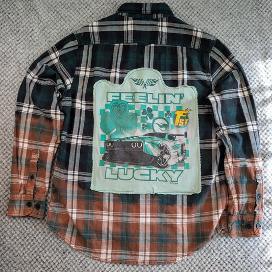 A long-sleeve plaid flannel shirt laid flat on a textured gray carpet, showing the back side. The shirt features a dark green and black plaid upper section that fades into rust brown and orange plaid toward the lower half.