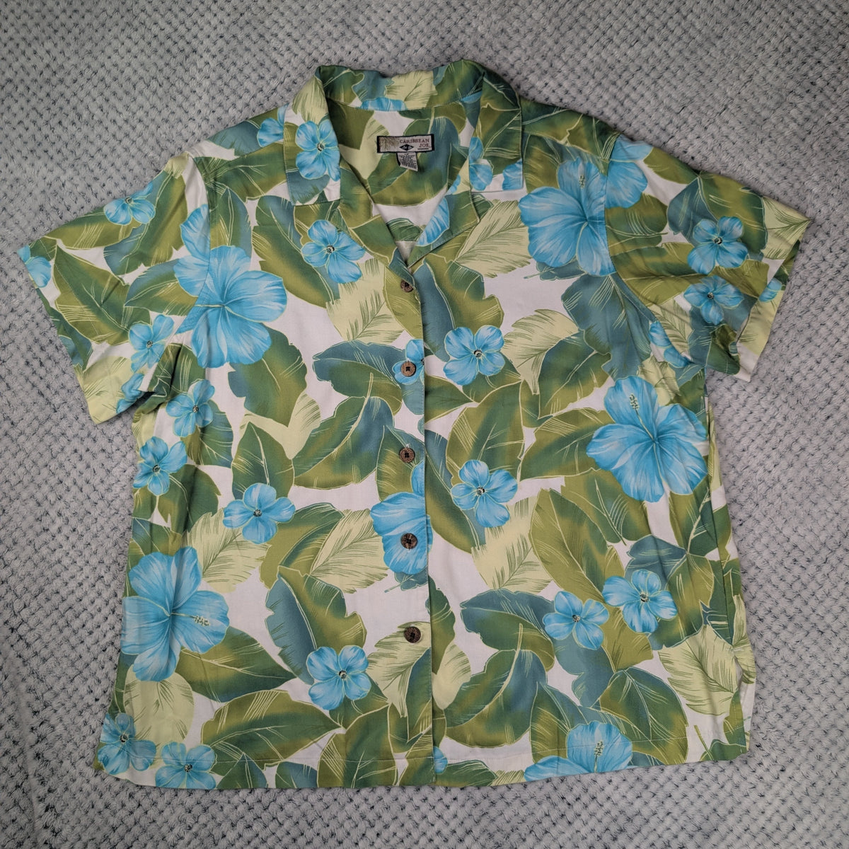A short-sleeve button-up camp shirt with a tropical floral print featuring blue hibiscus flowers and green leaves on a cream background, laid flat on a gray textured blanket.
