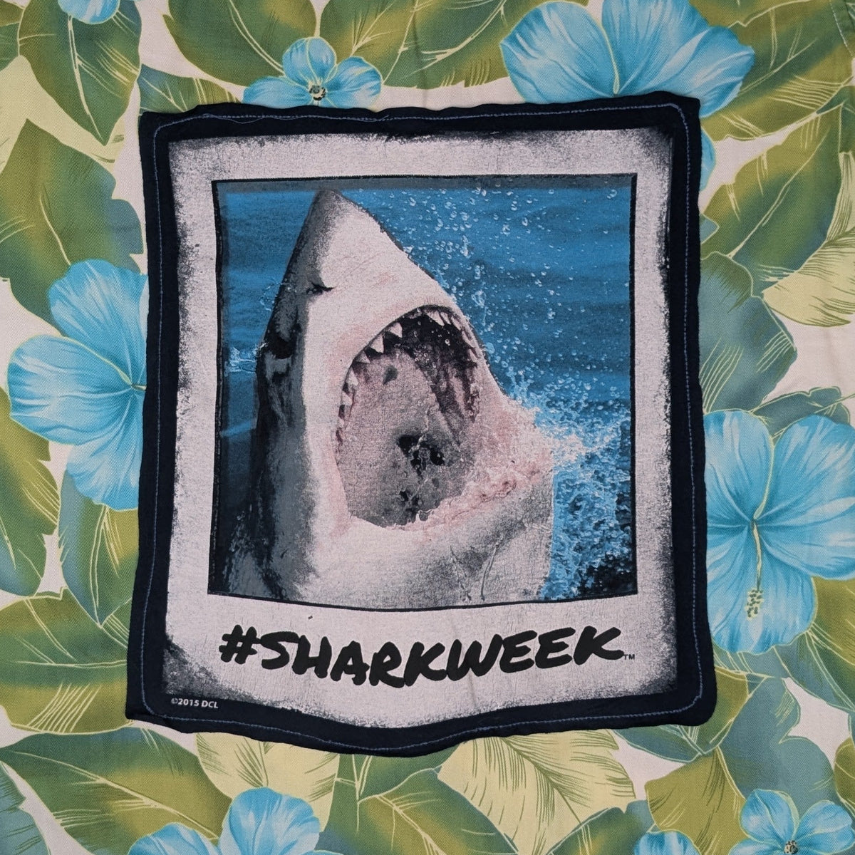 A short-sleeve Hawaiian-style shirt laid flat on a gray textured blanket. The shirt features a green and blue tropical floral print with a large square graphic on the front showing a shark in water and the text '#SHARKWEEK'.