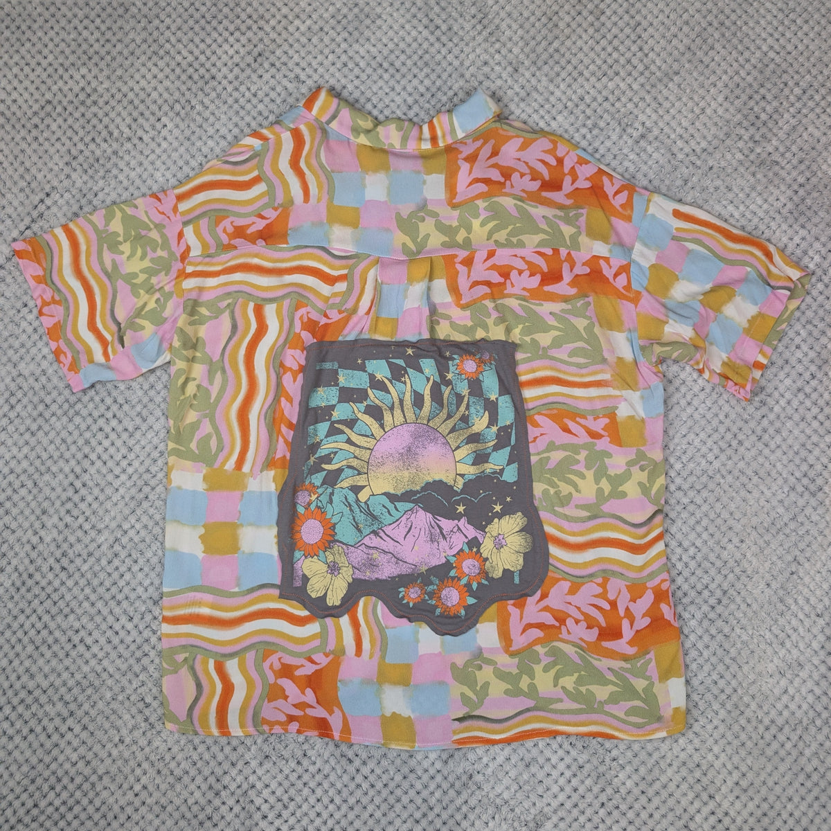A short-sleeve button-up shirt with a vibrant retro psychedelic pattern featuring wavy stripes, checkered blocks, and a central graphic print, laid flat on a gray textured background.