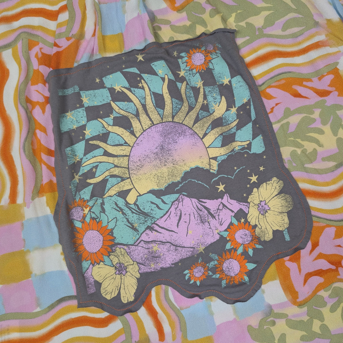 A retro psychedelic design featuring a sun, mountains, and flowers, laid flat on top of a multicolored patterned fabric background.