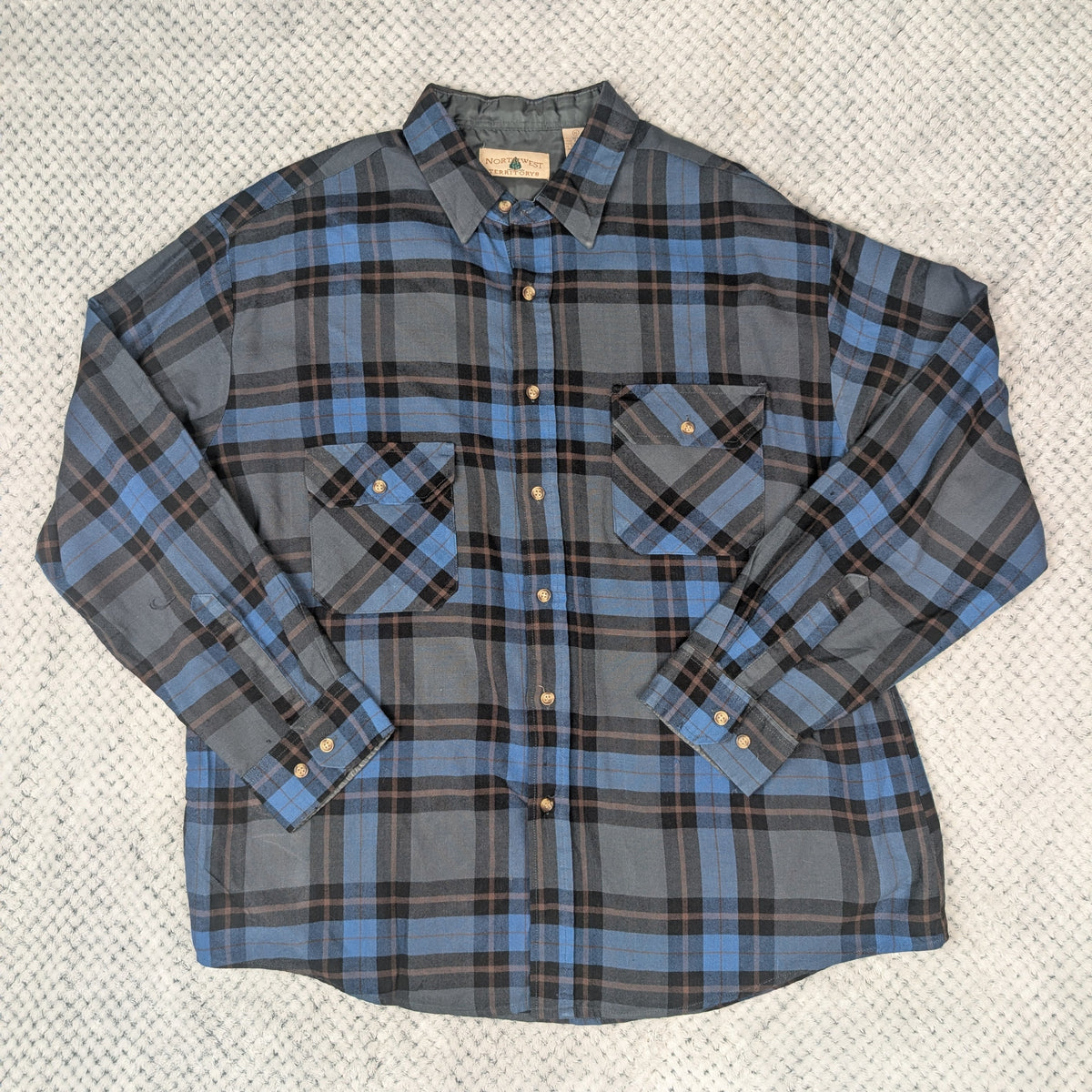 A long-sleeve plaid button-up shirt in blue, gray, and black, laid flat on a textured light gray knit blanket.