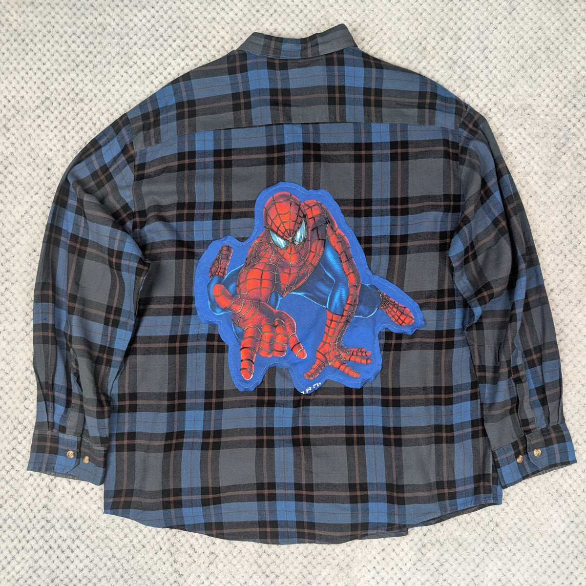 A long-sleeve plaid flannel shirt laid flat on a textured light gray surface. The shirt features a blue and black plaid pattern with a large Spider-Man graphic patch on the back, showing Spider-Man in red and blue with a blue outline.