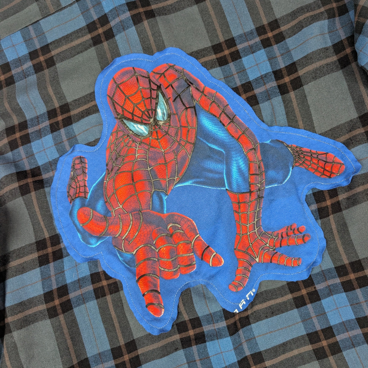 A Spider-Man themed patch or applique featuring a red and blue Spider-Man character design, placed on top of a blue and brown plaid fabric background.