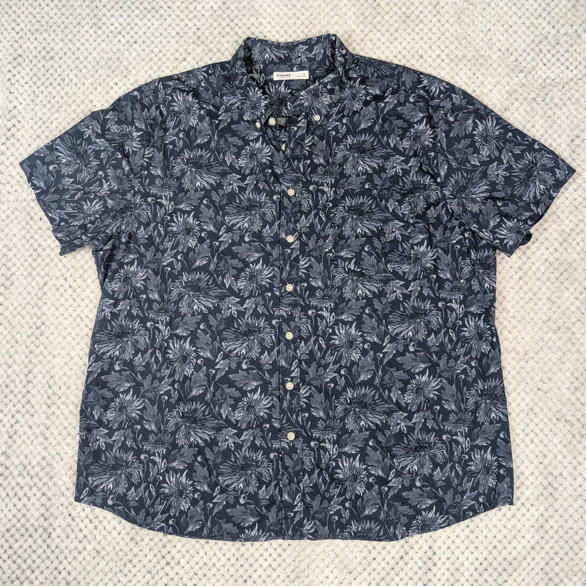 A short-sleeve button-up shirt with a dark navy blue and gray palm leaf pattern, laid flat on a textured light gray fabric background.