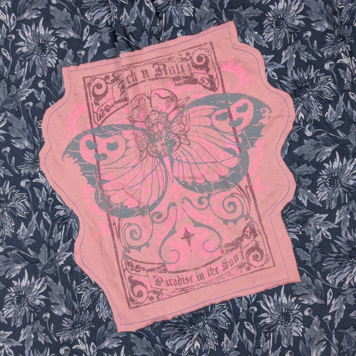 A pink fabric patch with a vintage-style butterfly illustration and decorative text, laid flat on a dark blue floral-patterned fabric background.