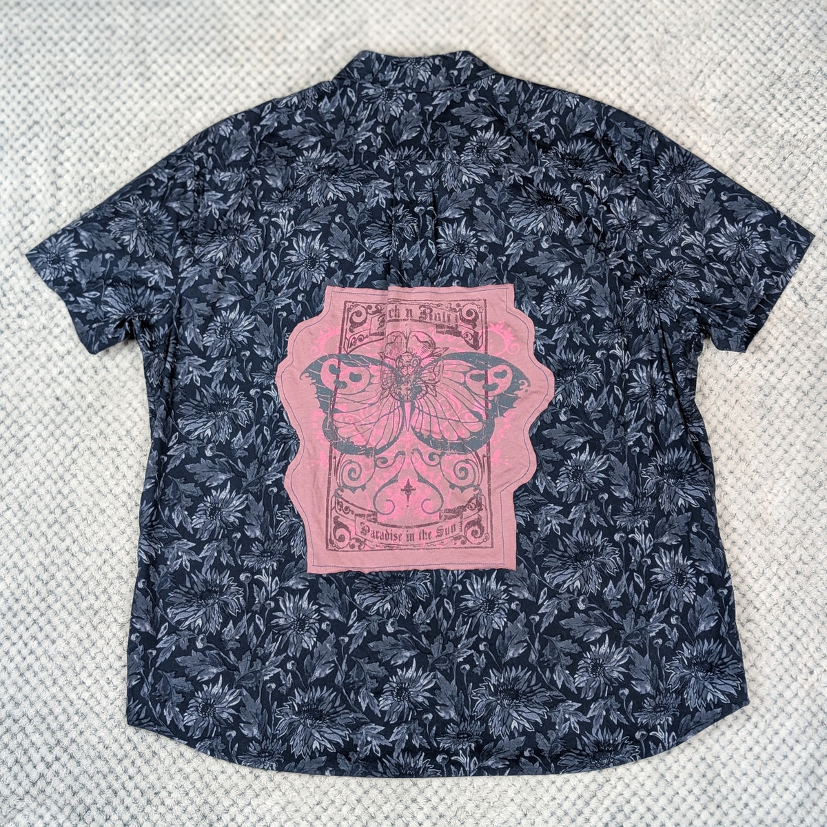 A short-sleeve button-up shirt laid flat on a textured light gray surface. The shirt features a dark navy blue and black tropical leaf pattern with a large pink graphic print on the back depicting a butterfly within a vintage playing card design.