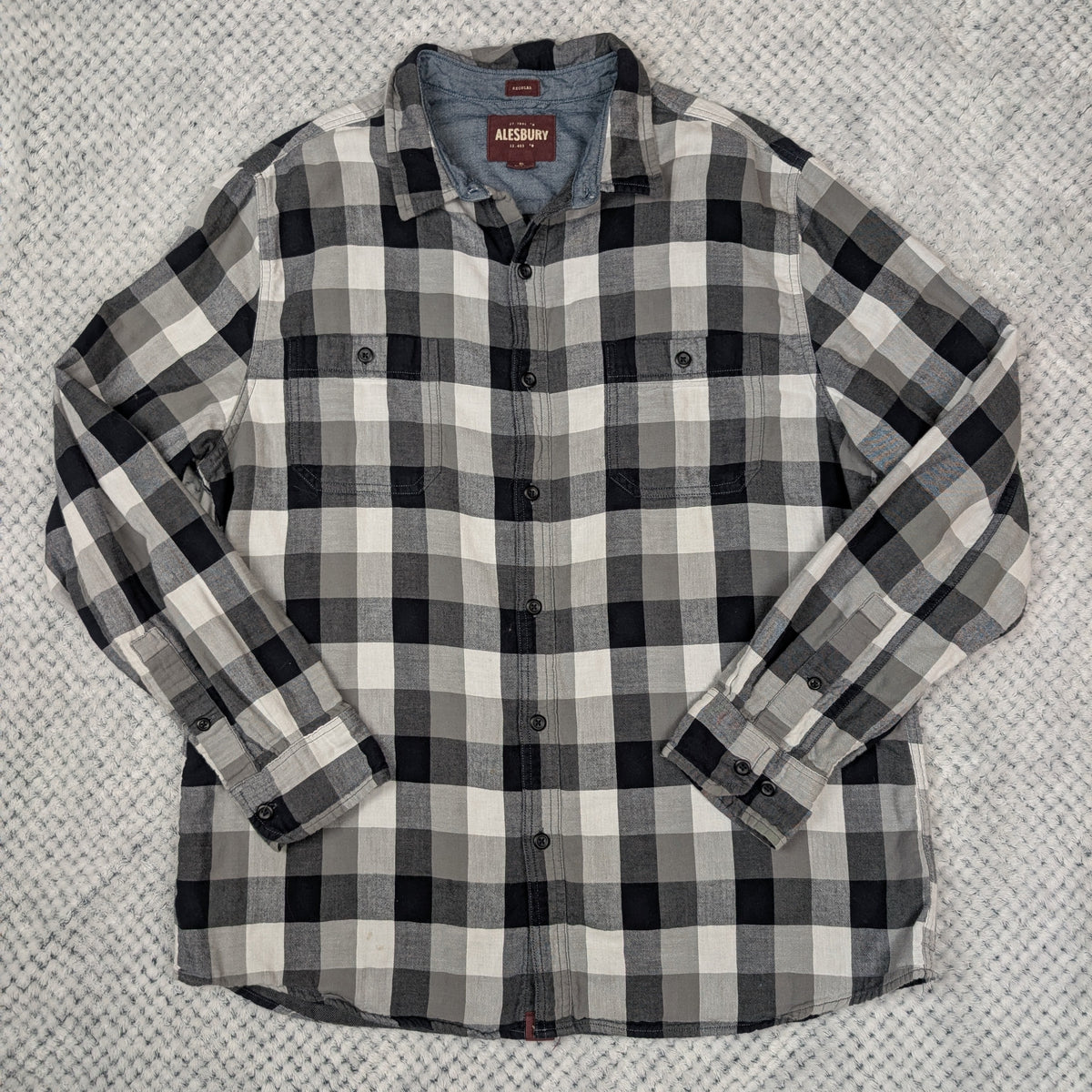 A long-sleeve plaid button-up shirt in black, white, and gray, laid flat on a textured gray blanket.
