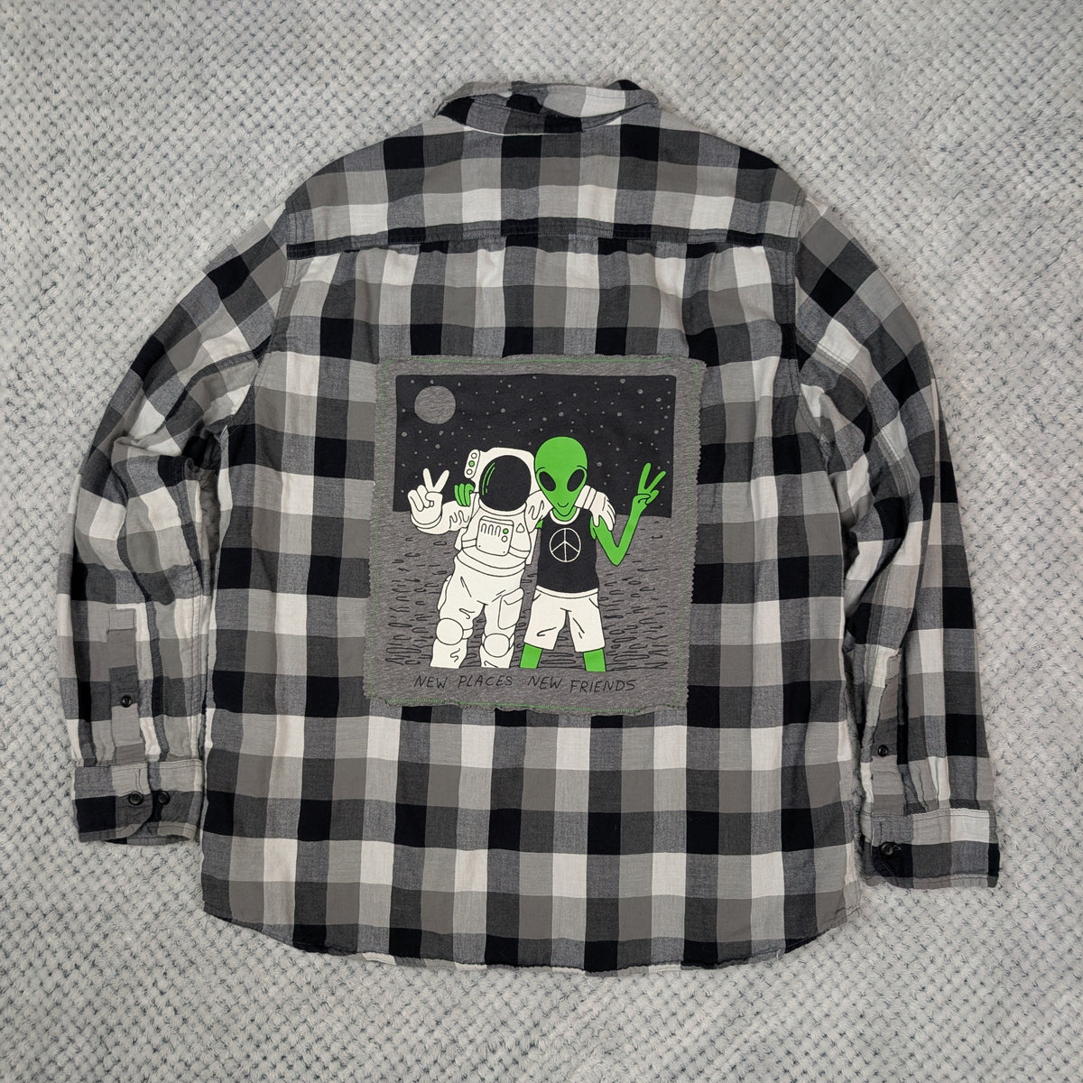 A black and white plaid long-sleeve flannel shirt laid flat on a textured gray background, featuring a central graphic print of an astronaut and a green alien on a gray square patch.