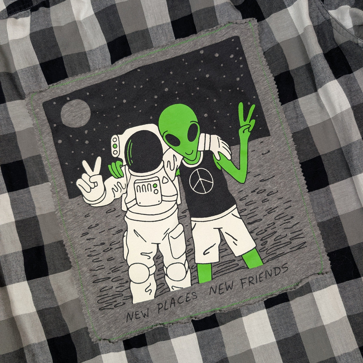 A square gray fabric patch with a retro space-themed illustration of an astronaut and a green alien on a black background, placed on a black and white plaid shirt. The patch has the text 'NEW PLACES NEW FRIENDS' at the bottom.