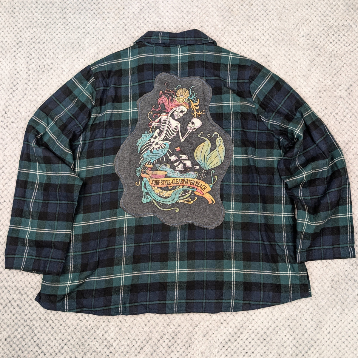 A long-sleeve plaid flannel shirt laid flat on a textured white blanket. The back of the shirt features a large graphic print of a skeleton with a mermaid tail.