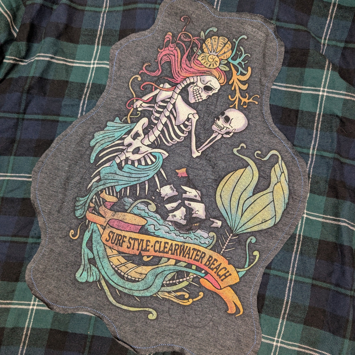 A dark gray heathered t-shirt featuring a large, colorful graphic of a skeleton mermaid with pink hair and a green tail, laid flat on a green and black plaid flannel shirt. The graphic includes the text 'SURF STYLE, CLEARWATER BEACH'.