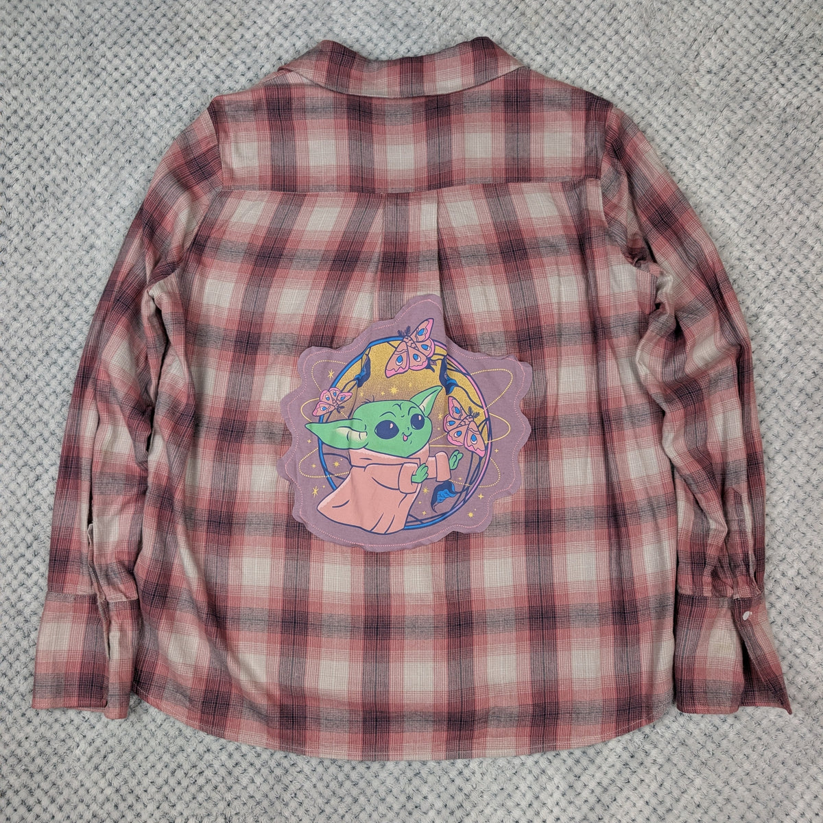 A pink and red plaid long-sleeve button-up shirt laid flat on a gray textured blanket, featuring a large graphic patch of Grogu (The Child) from Star Wars on the back.