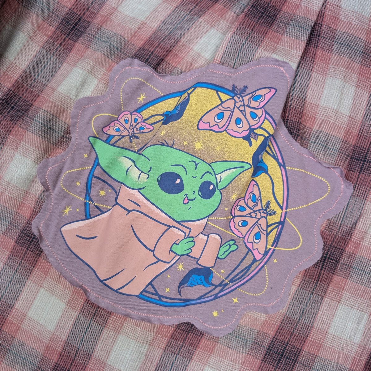 A scalloped patch featuring a cartoon green character in a beige robe surrounded by pink butterflies on a yellow and blue circular background, placed on a pink and gray plaid fabric surface.