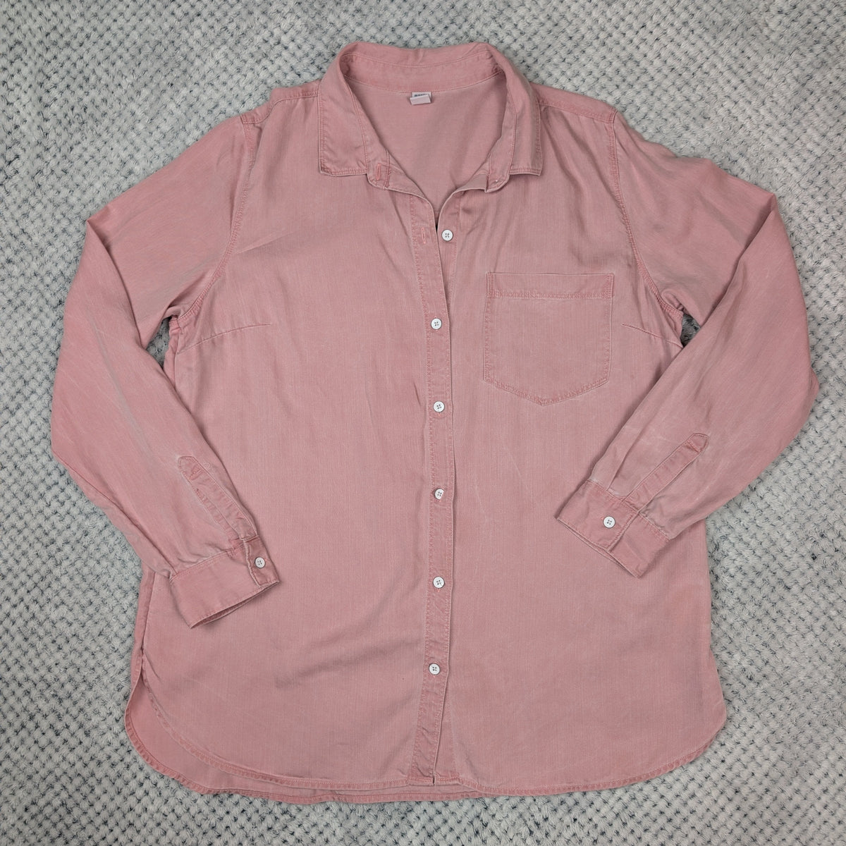 A dusty pink long-sleeve button-up shirt laid flat on a textured gray blanket. The shirt features a classic collar, a chest pocket, and white buttons down the front.