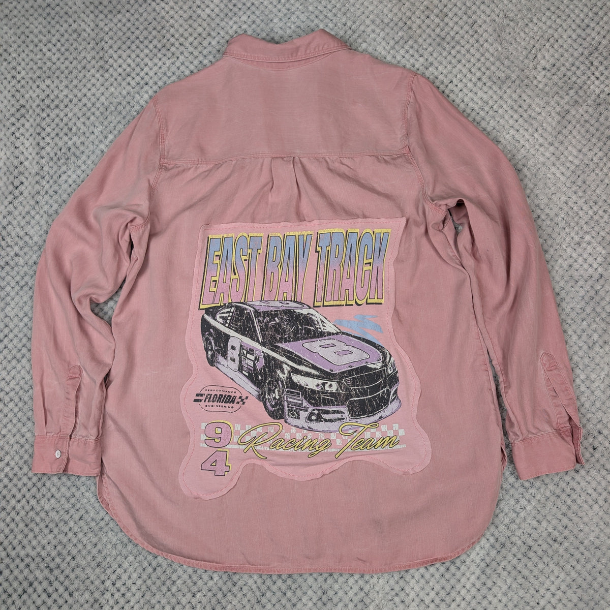 A pink long-sleeve button-up shirt laid flat on a gray textured fabric background, featuring a large graphic print on the back with a race car, 'East Bay Truck Racing' text, and '94'.
