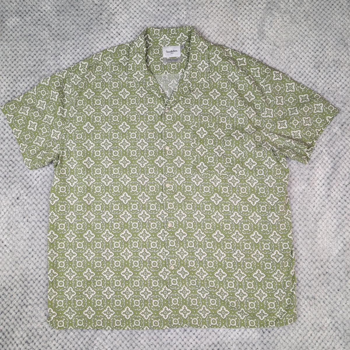 A short-sleeve button-up shirt with a light green and white geometric pattern, laid flat on a textured gray fabric background.