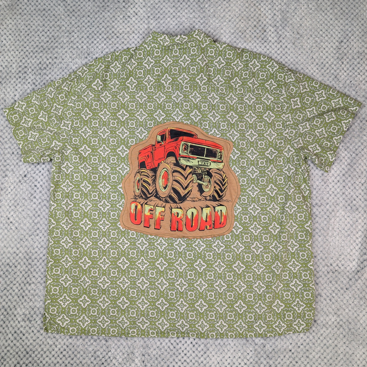 A short-sleeve button-up shirt laid flat on a textured gray background. The shirt features an all-over light olive green and white geometric pattern, with a large central graphic of a red monster truck and the words 'OFF ROAD' in red and yellow.
