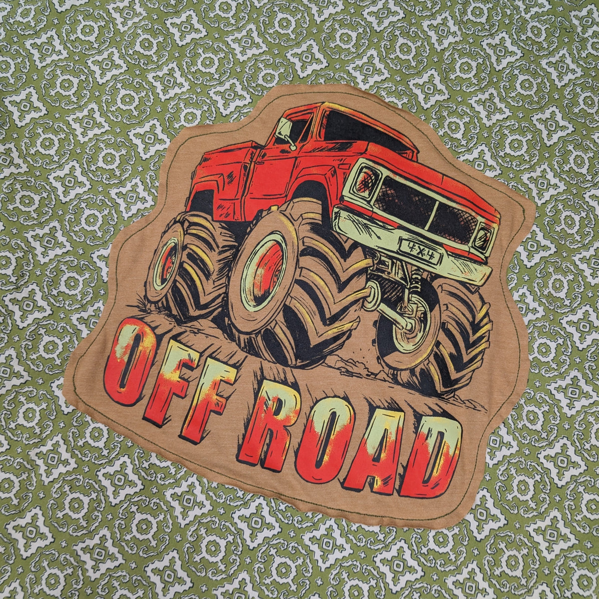 A die-cut graphic patch featuring a red monster truck with oversized tires and the words 'OFF ROAD' in red and cream lettering, placed on a green and white patterned fabric background.