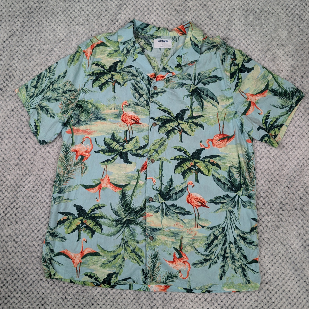 A short-sleeve button-up Hawaiian shirt with a tropical print featuring green palm trees, pink flamingos, and a light blue-green background, laid flat on a gray textured blanket.