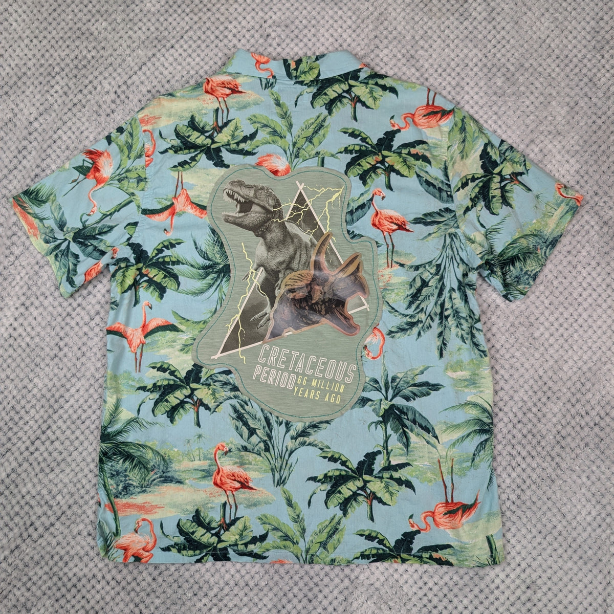 A short-sleeve button-up shirt with a light blue tropical print featuring green palm trees, pink flamingos, and a large dinosaur graphic on the front, laid flat on a gray textured background.