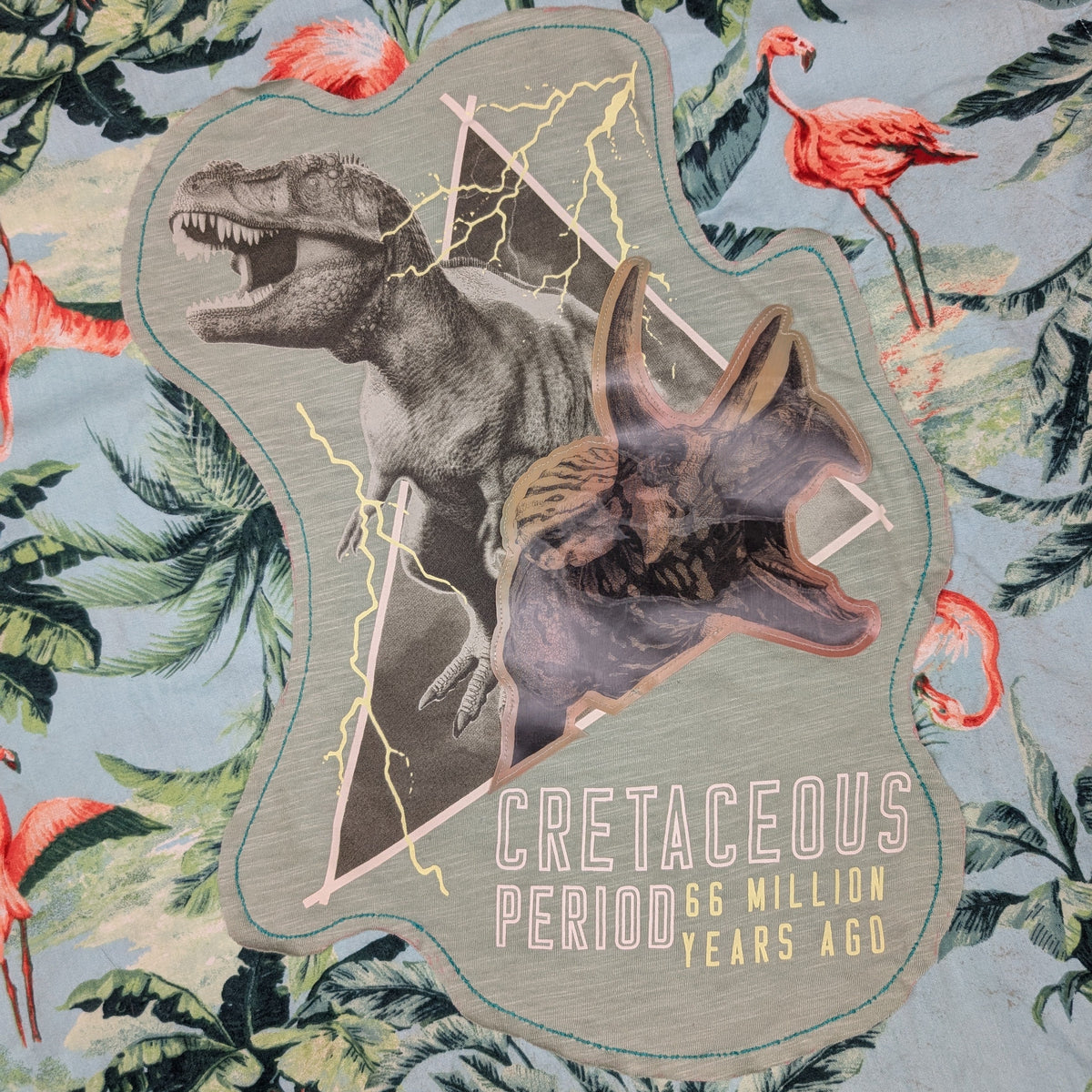 A close-up of a fabric surface featuring a tropical print with green palm leaves, pink flamingos, and a large central dinosaur collage graphic showing a T-rex and a pterosaur with lightning and the text 'CRETACEOUS PERIOD 66 MILLION YEARS AGO'.