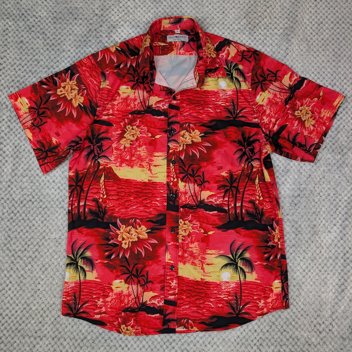 A short-sleeve button-up Hawaiian shirt with a vibrant red tropical sunset print, laid flat on a gray textured carpet.
