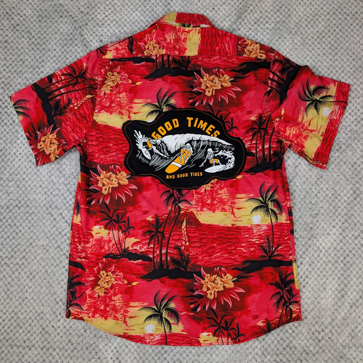 A red Hawaiian short-sleeve button-up shirt with a tropical sunset print, laid flat on a gray textured background. The shirt features a large black patch on the back with the text 'GOOD TIMES' and 'SEA LIFE TOYS' around a shark graphic.