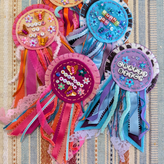 Five colorful handmade ribbon rosettes with lace and sequin embellishments, featuring text like 'hobby hoe' and 'Crafty Bitch,' arranged on a striped fabric background.