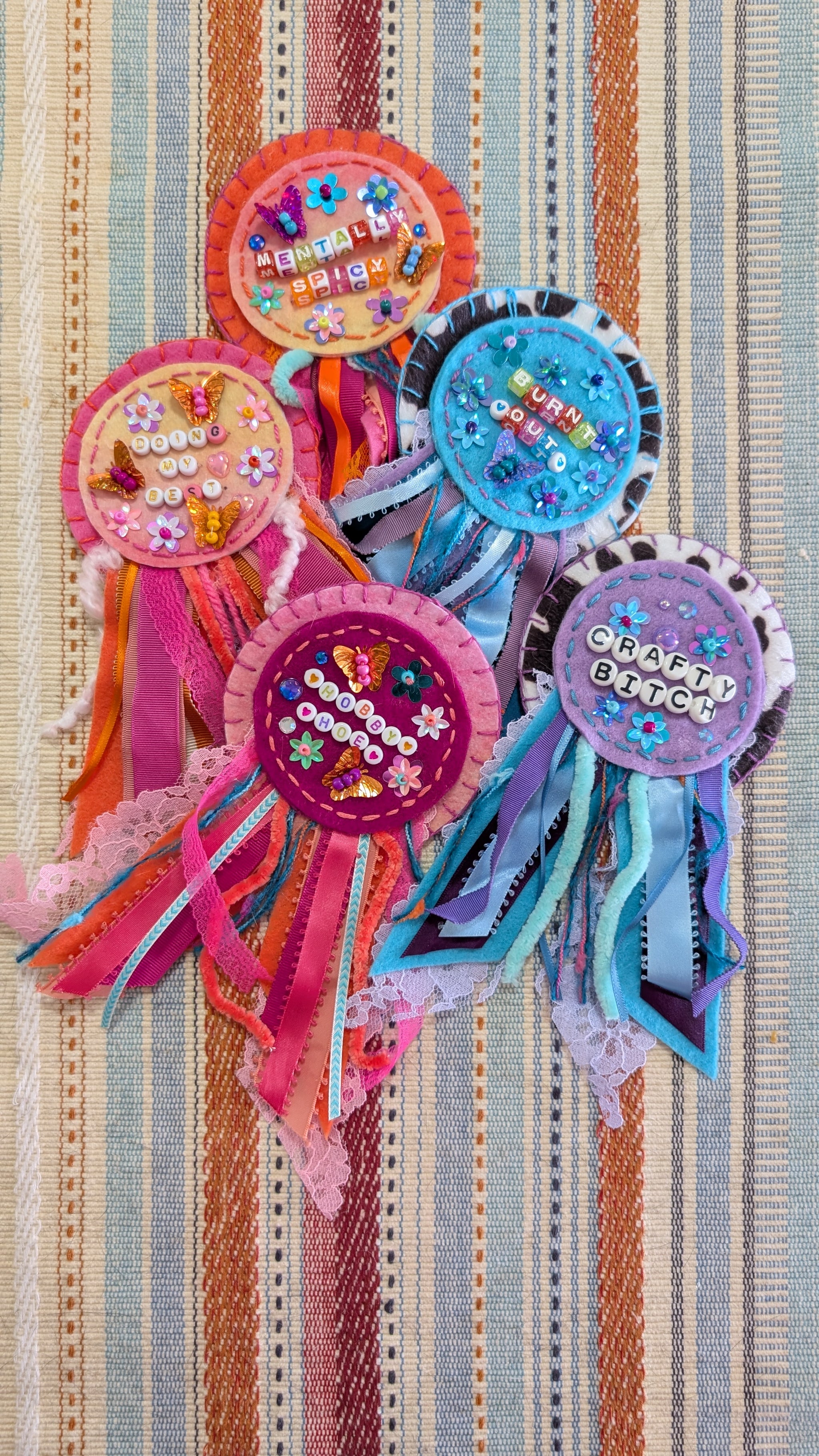 Five colorful handmade ribbon rosettes with lace and sequin embellishments, featuring text like 'hobby hoe' and 'Crafty Bitch,' arranged on a striped fabric background.