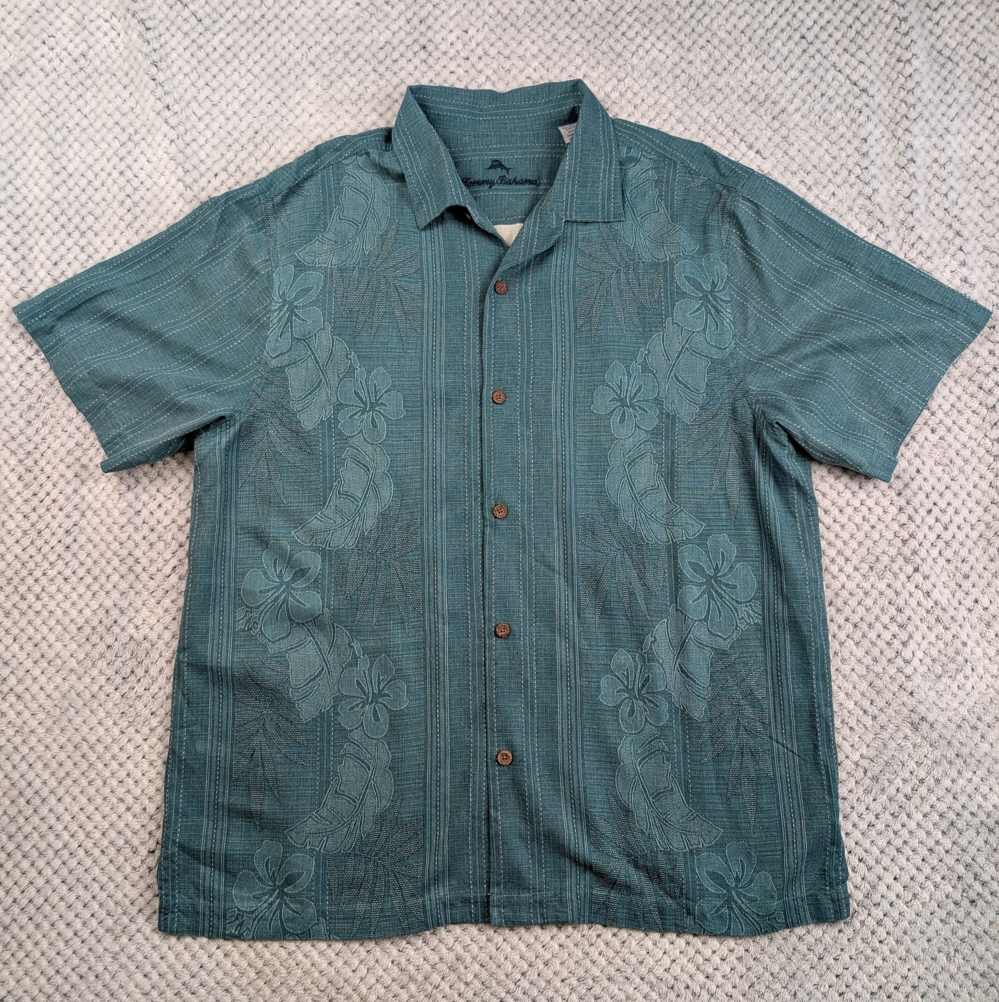 A teal short-sleeve button-up shirt with a subtle floral jacquard pattern, laid flat on a textured beige blanket.